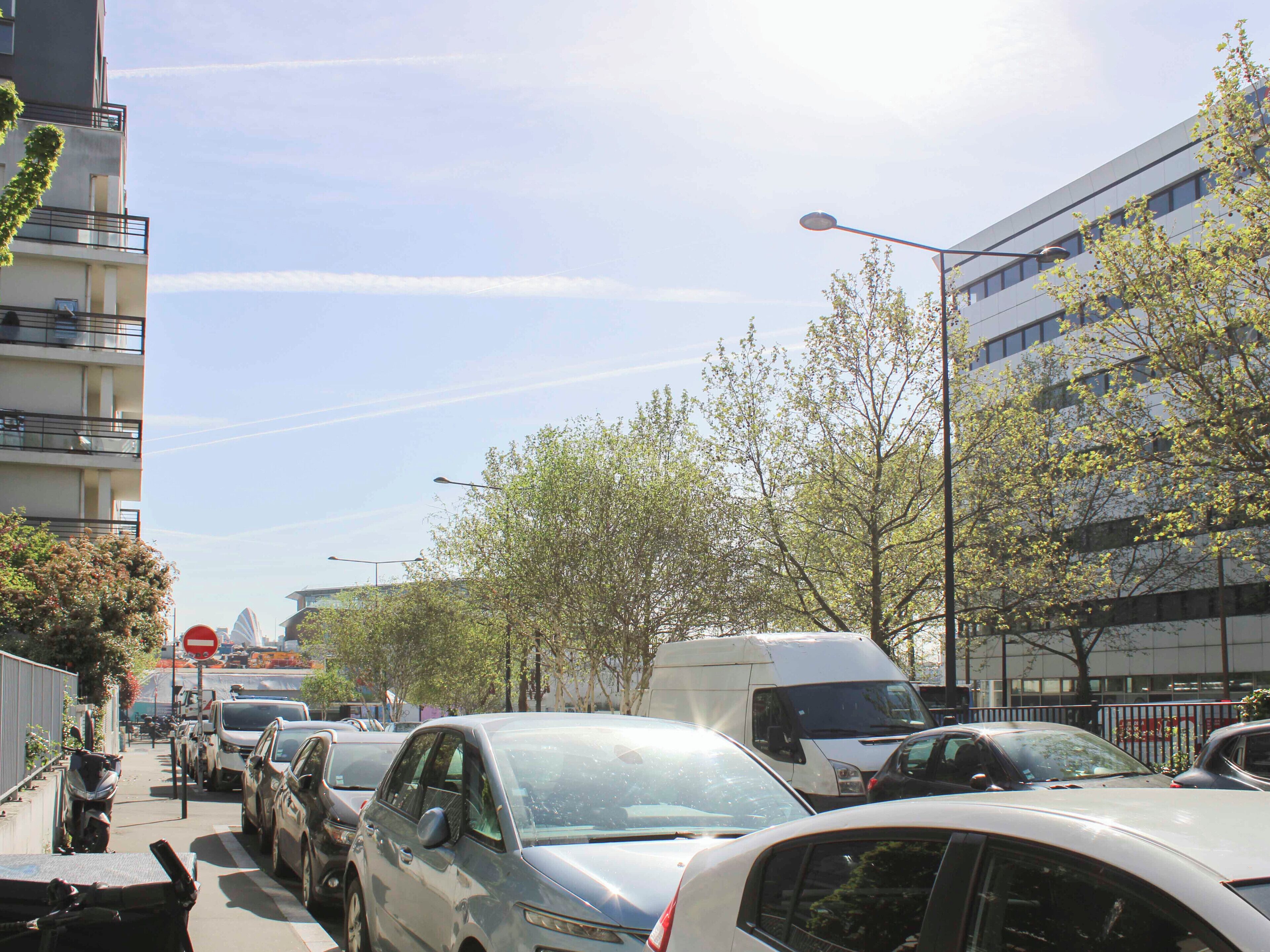 A street lined with parked cars and buildings, a clear sky, and a no entry sign.