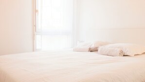 Un llit amb roba de llit blanca en una habitació de parets blanques i una finestra.