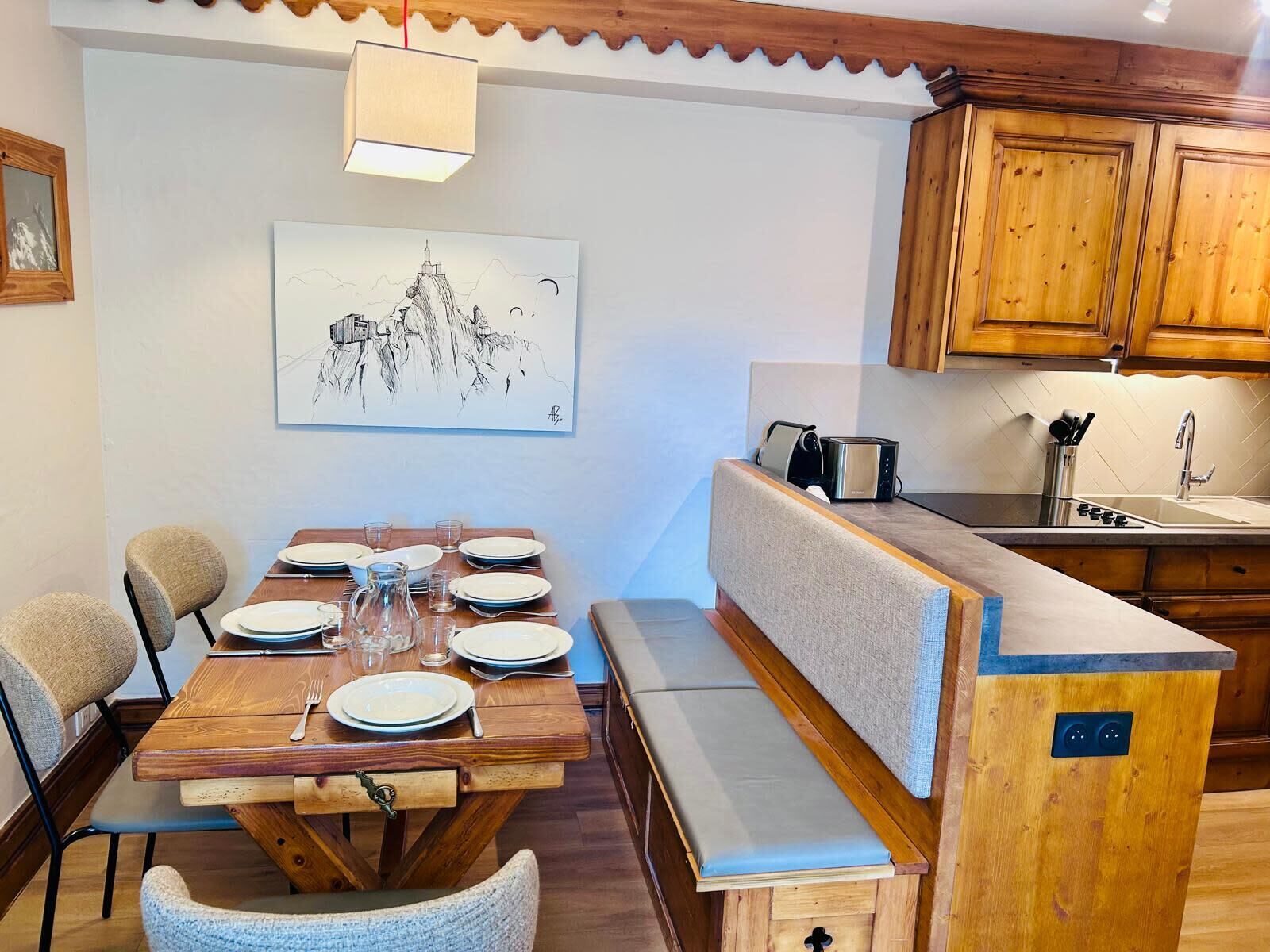 Una mesa de comedor de madera con platos y vasos, un banco y una zona de cocina con armarios de madera y un lavamanos.