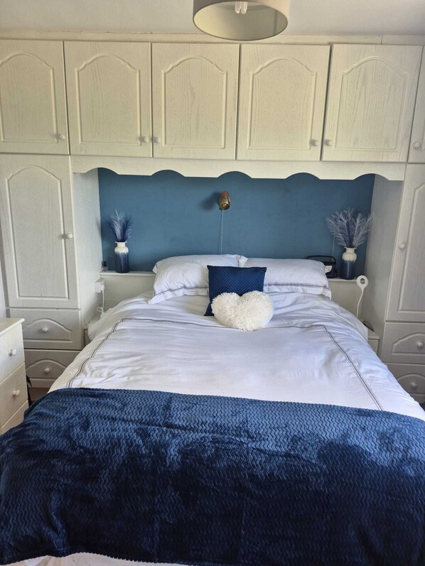 Une chambre à coucher avec un lit, des tables de chevet blanches, une commode, un mur d’accent bleu et un luminaire au plafond.