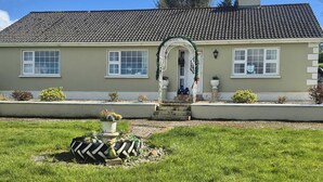 Une maison à deux étages, avec un toit recouvert de tuiles, un porche à l’avant doté d’une balustrade blanche et un petit jardin avec un chemin menant à l’entrée.