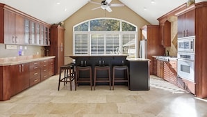 A kitchen with a central island, bar stools, wooden cabinets, a large window, and a ceiling fan.