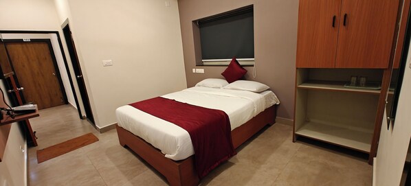 A bedroom with a bed, a wooden wardrobe, a window with a blind, and a door with a number. - AKSHAYA COMFORTS (Visakhapatnam)