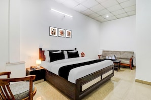 A bedroom with a large bed, a bedside table with a lamp, a chair, a sofa, and three framed pictures on the wall. - Deluxe Bhagwan Guest House (Dehradun)