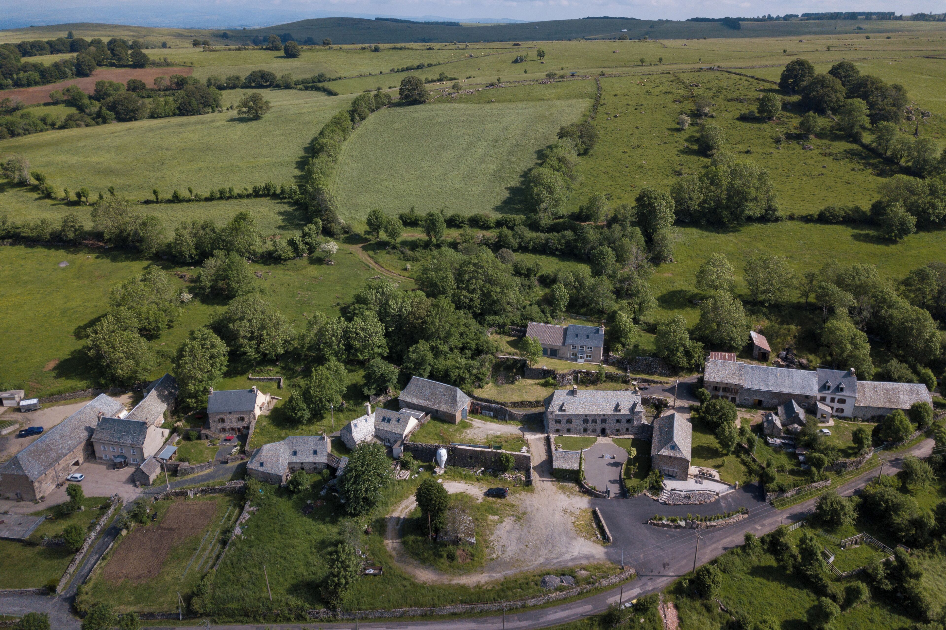 Vista aérea de uma aldeia rural com edifícios de pedra, um pátio central e campos ao redor.
