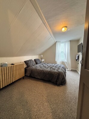Une chambre avec un plafond en pente, un lit recouvert d’une couverture sombre, une table de chevet en bois, une lampe, un téléviseur et une fenêtre avec des rideaux.