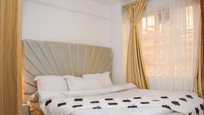 A neatly made bed with a headboard, a window with sheer curtains, and a wooden floor. - Serene 1-bedroom apartment in Nairobi perfect for your stay (Nairobi)