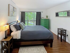Ein Schlafzimmer mit einem großen Bett, einem Nachttisch, einer Kommode, einem kleinen Tisch und einem Fenster mit Vorhängen.