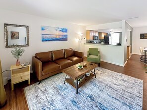 A living room with a brown leather sofa, a wooden coffee table, a green armchair, a painting of a sunset, and a kitchen area in the background. - Brand New! Serene Vista View 2-BR 2BA Condo: 1.8 ml to UF & Downtown/Pool & Gym (Gainesville)