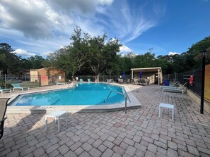 Ein rechteckiger Pool mit einer Fliesenveranda, umgeben von Liegestühlen und Sonnenschirmen, mit Bäumen und einem Zaun im Hintergrund.