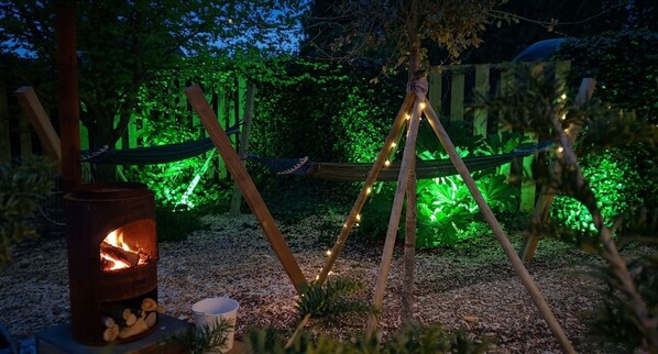 Un cadre extérieur rustique avec un poêle à bois, un hamac et des guirlandes lumineuses.