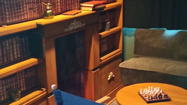 Quarto com estante de livros, uma cadeira azul, uma mesinha redonda com um tabuleiro de xadrez e um sofá.