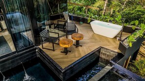 Une terrasse extérieure moderne, dotée d’une paroi vitrée, d’un plancher en bois, d’une baignoire blanche et entourée de verdure.