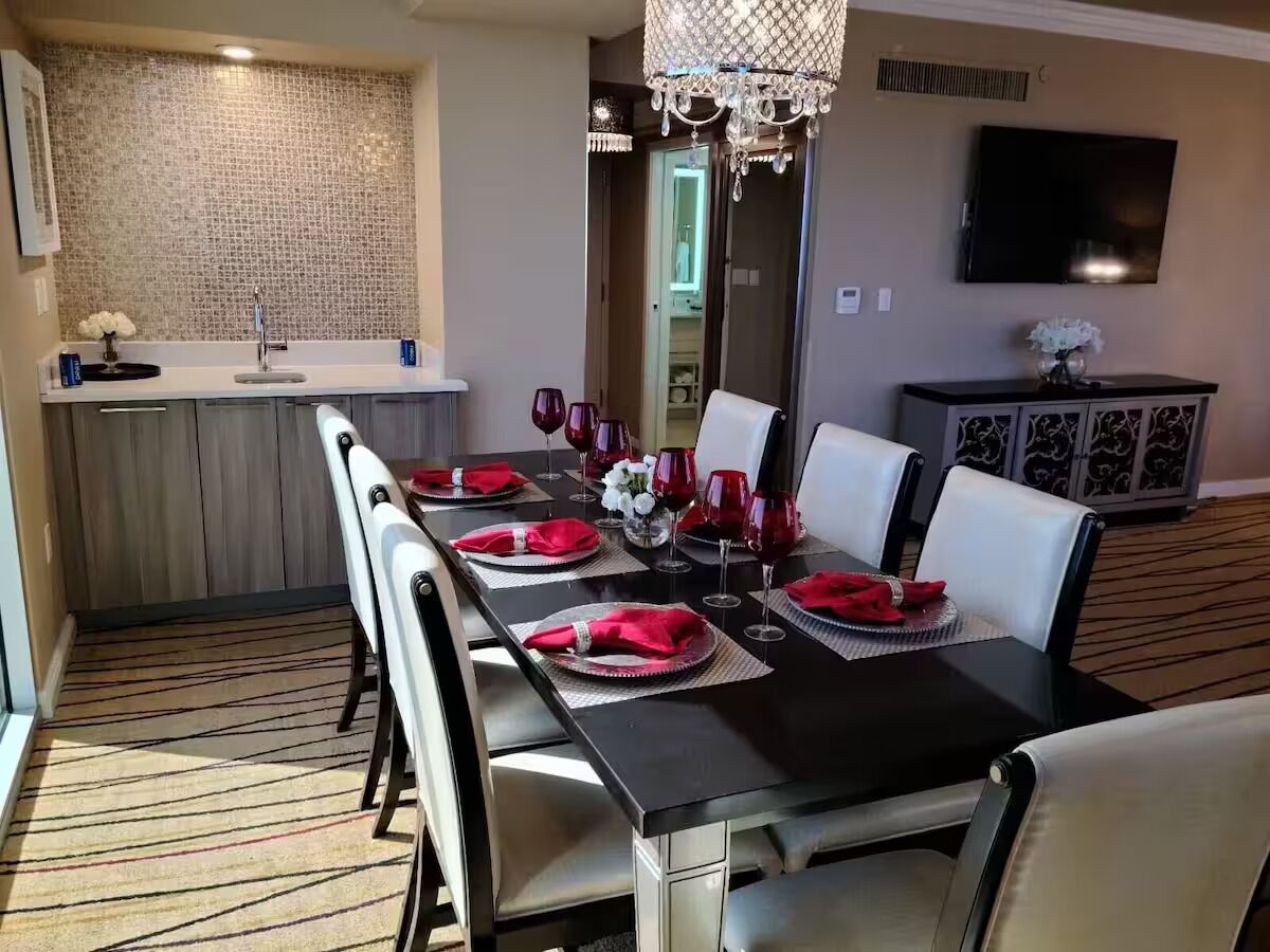 Comedor con una mesa negra dispuesta para seis personas, sillas blancas, una araña, televisor en la pared y una zona de cocina con salpicadero de azulejos.