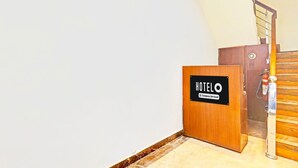 Uma área de recepção de hotel com um balcão de madeira identificado como 'HOTELO Company-Serviced', um extintor de incêndio e uma escada de madeira.