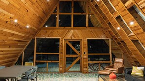 Interior de madera con techo alto, ventanas amplias y zona de estar con mesa y sillas.