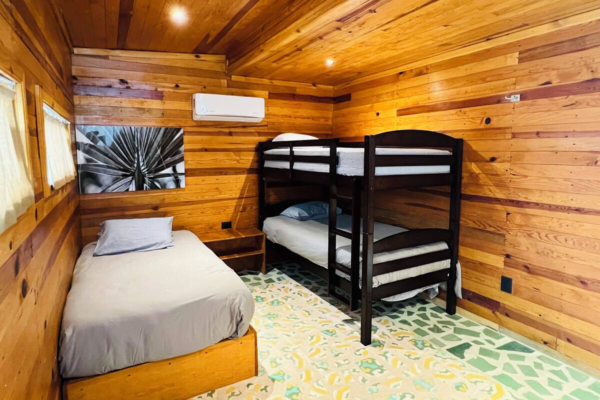 Una habitación de cabaña de madera con literas, una cama, aire acondicionado montado en la pared y suelo estampado.