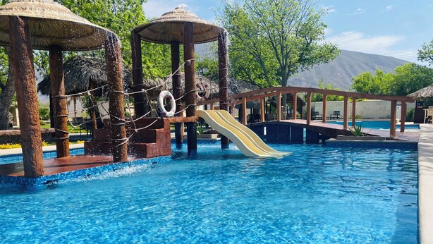 Una piscina con tobogán, columpios de cuerda y estructuras de techo de paja.