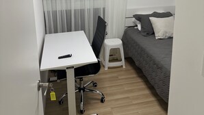 Et lille, moderne værelse med et hvidt skrivebord, en sort kontorstol, en grå seng med puder, en hvid skammel og et stort vindue med lette gardiner.
