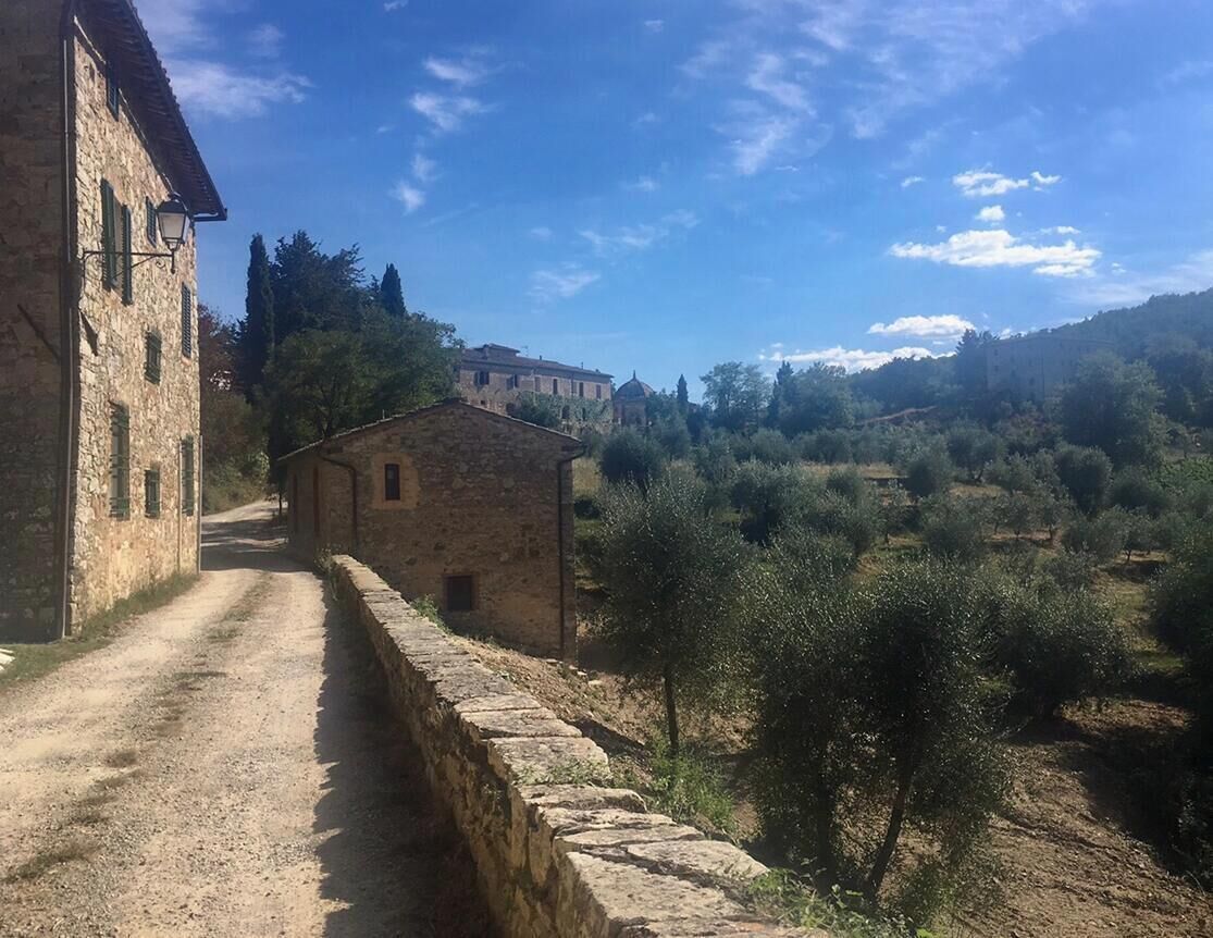 A stone building with a dirt path leading to a small house, surrounded by olive trees and hills.