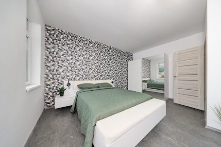 A modern bedroom with a geometric-patterned wall, a bed with a green bedspread, a bedside table with a plant, and a wardrobe.