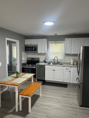 Una cocina moderna con armarios blancos, electrodomésticos de acero inoxidable, una encimera de granito y una zona de comedor con mesa y banco de madera.