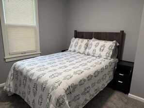 Un dormitorio con cama, mesita de noche, ventana con persianas y suelo alfombrado.