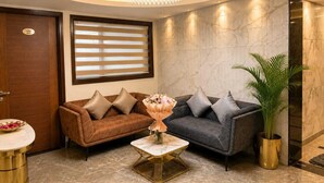 A lobby with a brown and grey sofa set, a gold-framed coffee table, a chandelier, and a potted plant. - Sukhvan Residency (Gurugram)