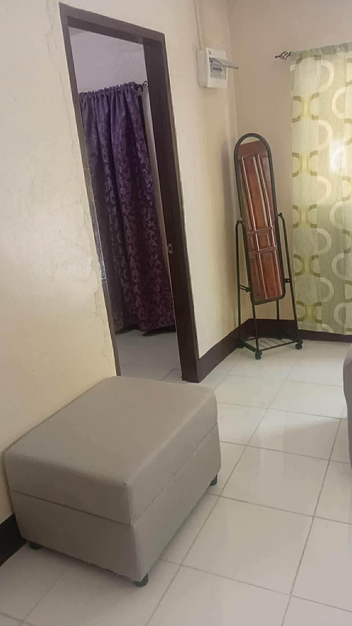 Une pièce avec un sol carrelé de carreaux blancs, un pouf gris, une porte ornée d’un rideau violet et un radiateur portatif.