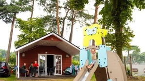 Une aire de jeux avec un toboggan et une structure d’escalade, entourée d’arbres et bordée d’un bâtiment au toit rouge.