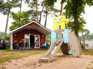 En legeplads med rutchebane og klatrestruktur, omgivet af træer og en bygning med rødt tag.