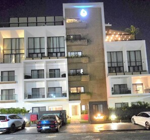 Un hotel de varios pisos con balcones, un elemento decorativo luminoso en el centro y coches aparcados.