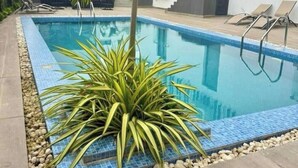 Piscina com borda azulejada, cercada por pedras e plantas, com espreguiçadeiras e um edifício moderno ao fundo.