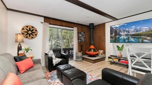A living room with a sofa, ottoman, fireplace, and a large window with a view of trees. - Rocky’s Riverside Retreat - heated pool, river access (Estes Park)