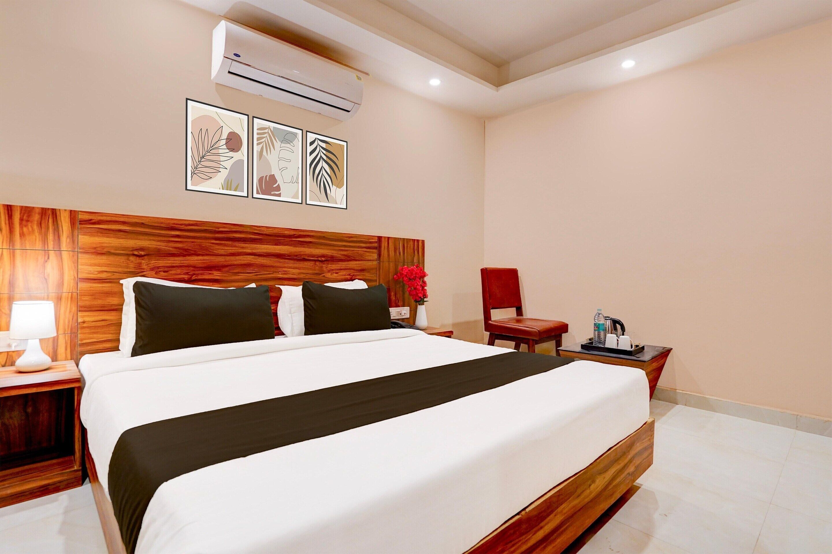 A hotel room with a wooden headboard, a large bed, a nightstand with a lamp, a red chair, and a small table with a bottle and a cup.