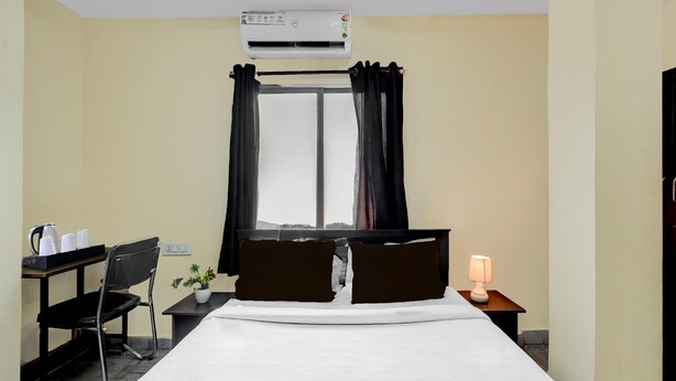 A hotel room with a bed, a desk with a chair, a small table with a lamp, an air conditioner, and a window with curtains. - Deluxecollection O Kothapet Near Omini Hospital (Hyderabad)