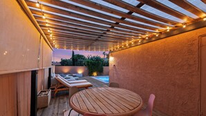 Een houten terras met een ronde tafel en stoelen, een zwembad en een pergola met lichtjes.