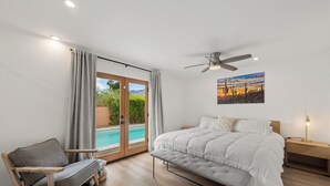 Een slaapkamer met een bed, een bankje, een stoel, een plafondventilator, een nachtkastje en een schilderij.