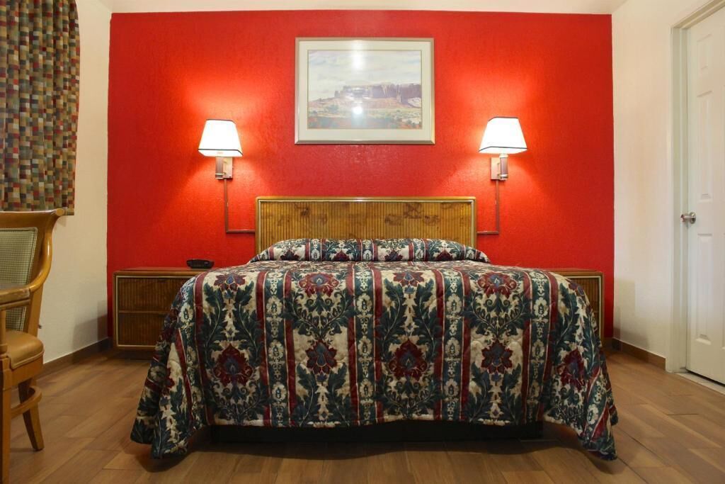 A bedroom with a red wall, two bedside lamps, a bed with a patterned bedspread, a wooden headboard, and a framed picture above the bed.