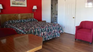 Una habitación con cama, una mesa de madera, un sillón rojo, una puerta blanca y un baño visible a través de una puerta abierta.