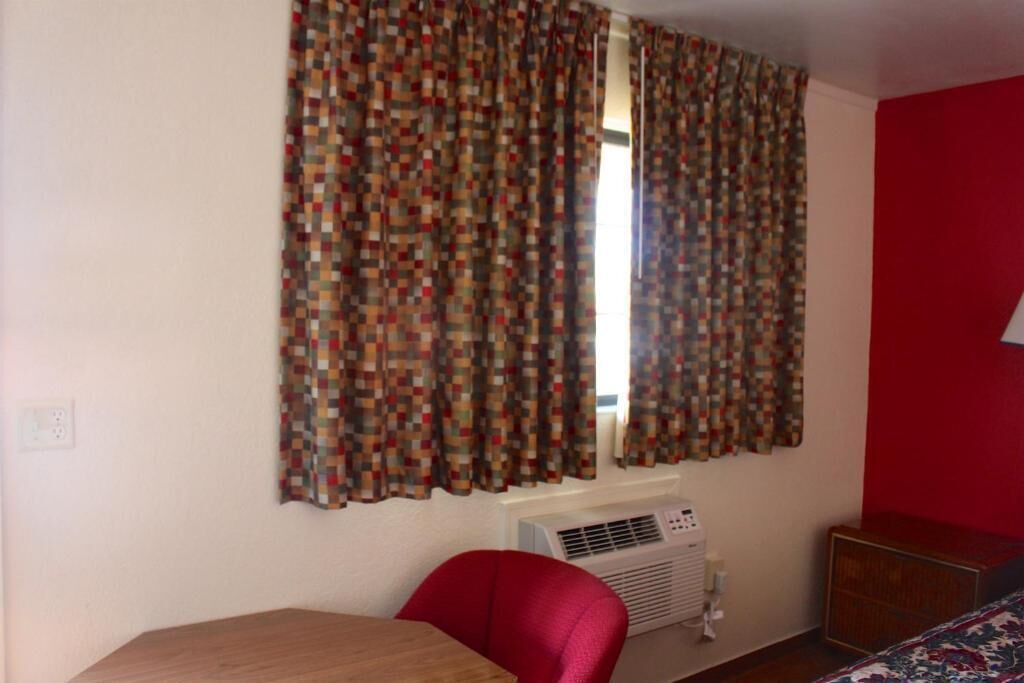 A room with a window covered by patterned curtains, a red chair, a wooden table, and a wall-mounted air conditioning unit.