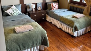 Une chambre avec deux lits, un plancher en bois, une table de chevet, une lampe, un tableau encadré et une fenêtre avec des rideaux.