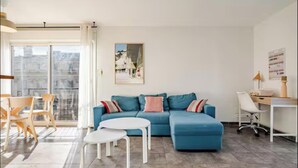 Een woonkamer met een blauwe bank, een witte salontafel, een bureau met stoel, en uitzicht op de stad door het raam.
