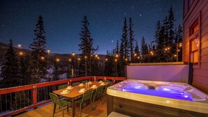 Une terrasse en bois avec un jacuzzi, une table et des chaises d’extérieur, le tout entouré d’arbres et illuminé de guirlandes lumineuses.