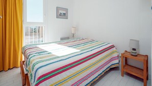 A bedroom with a bed, a window with yellow curtains, a bedside table with a lamp, and a wall-mounted picture. - Serene Port-side Living in Agde (Agde)