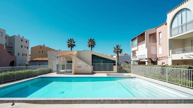A swimming pool with a clear blue water, surrounded by a white fence, and buildings in the background. - Serene Port-side Living in Agde (Agde)