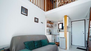 A compact living space with a sofa, a small kitchen area, and a door leading to a staircase. - Serene Port-side Living in Agde (Agde)