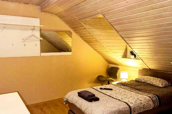 Ein kleines, gemütliches Schlafzimmer im Dachgeschoss mit schrägen Wänden, einem Einzelbett, einem Sessel, einem Schreibtisch und einem Kleiderhaken.
