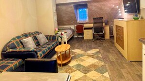 A compact living space with a sofa, a small round table, a desk with a chair, a TV, and a window. - Gemutliche Wohnung in Konz an der Mosel (Konz)