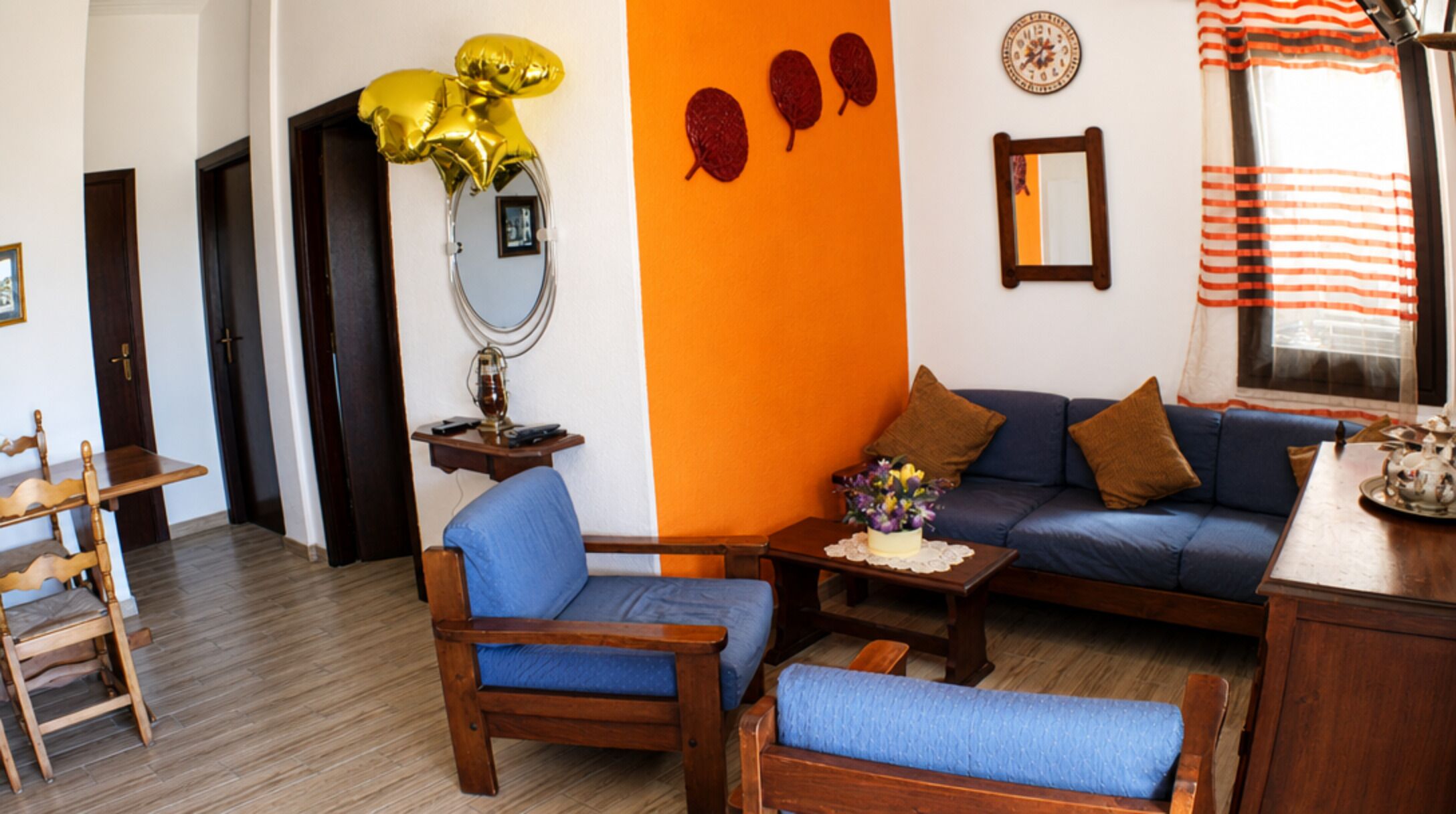 A living room with a blue sofa, wooden chairs, a small table, a mirror, and decorative items on the wall.